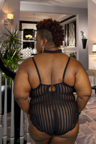 Rear view of plus size black lace bodysuit on curvy model with striped sheer panels and lace trim in a home interior